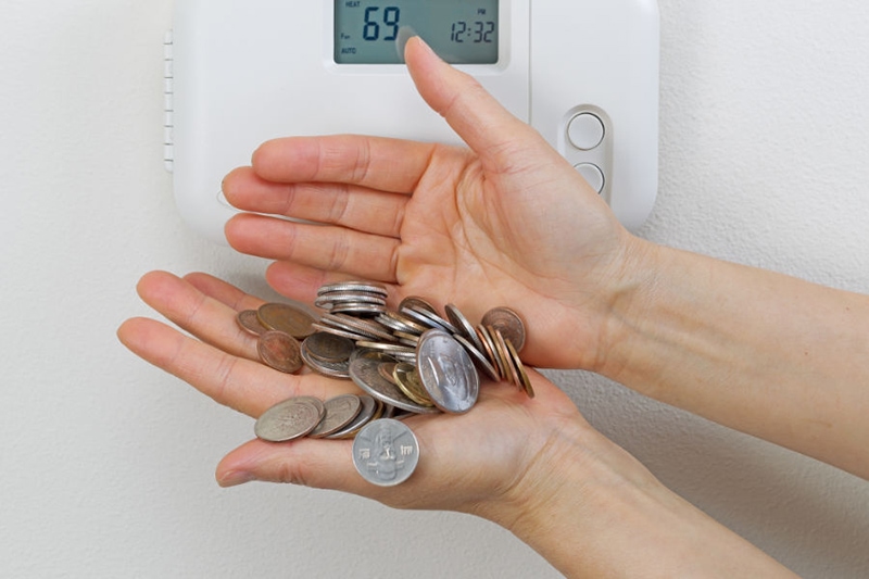 Hand holding quarters next to thermostat, 5 Ways to Save Money on Your Air Conditioning & Summer Energy Bill