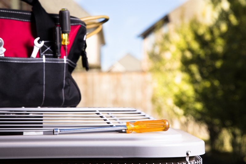 Bag of repairman's work tools, screwdriver on top of air conditioner unit outside brick home. Service industry, working class.