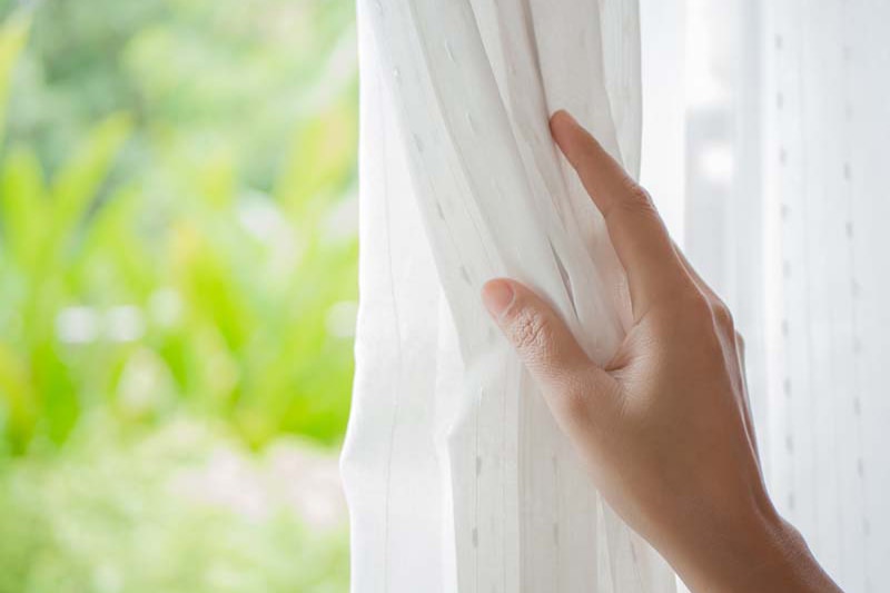 Woman's hand opening curtains in the bedroom with natural light and garden background.